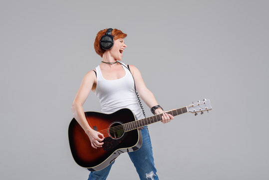 Young woman musician with an acoustic   guitar in hand and in headphones on a gray background. He laughs and plays rock and roll loudly. 