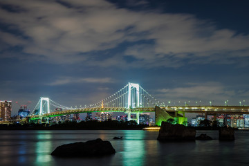 Rainbow bridge at night shot from Odaiba island, Tokyo, Japan. Dark rocks in the front stick out from the water, and scyscrapers illuminated with lights are in the background