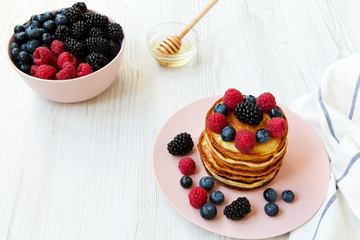 Pancakes with berries and honey on a pink plate over white wooden background, side view. Closeup.