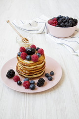 Pancakes with berries and honey on a pink plate, side view. White wooden table.