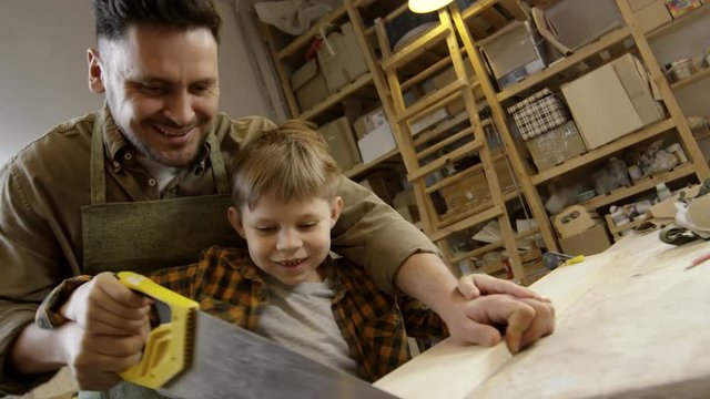 Medium shot of happy father and curious little boy sawing piece of plank in carpentry workshop