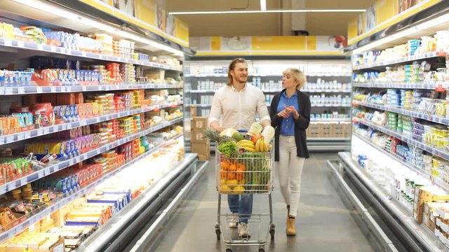 Caucasian cheerful male and female shoppers push cart with grocery products in supermarket aisle