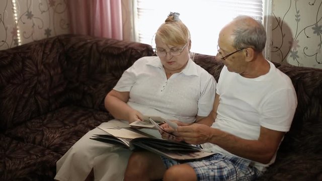 an elderly couple looks at their photos sitting at home on the couch. husband and wife with glasses watching his album.