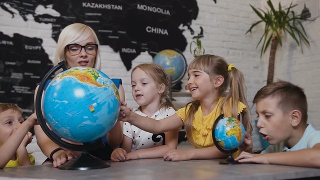 Beautiful pupils and teacher looks at globes in geography classroom on the lesson at the elementary school. Boys, girls and their young teacher study the map of the world using the globe and