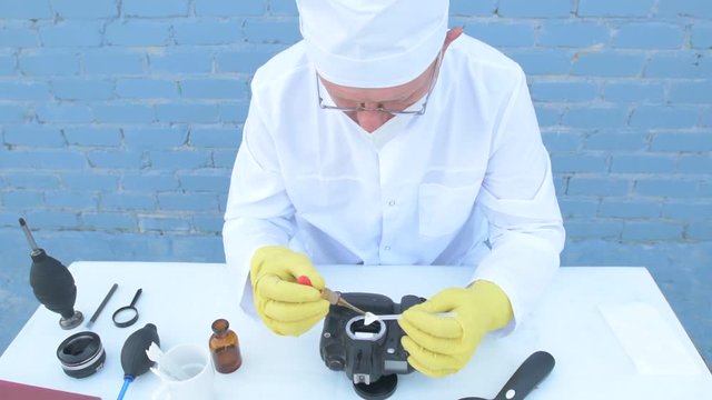 Elderly solid person in white dressing gown, cap, medical mask, glasses cleans matrix of digital camera with special means. Lens, sensor, matrix, cleaning wands, magnifier, isopropyl alcohol.