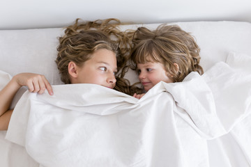 portrait of two young beautiful kids resting on bed, smiling and covering with sheets. white room. Family lifestyle indoors.