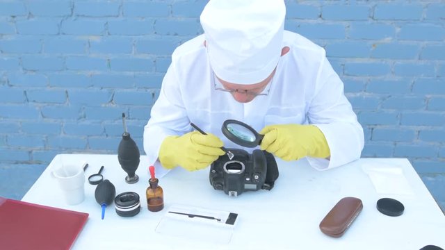 Elderly solid person in white dressing gown, cap, medical mask, glasses cleans matrix of digital camera with special means. Lens, sensor, matrix, cleaning wands, magnifier, isopropyl alcohol.
