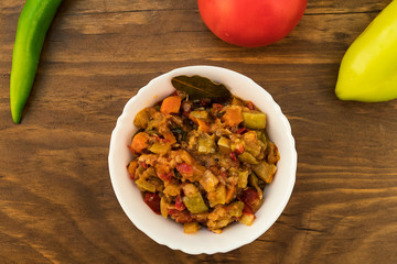 Stewed mixture of vegetables-zucchini, tomatoes, carrots, sweet peppers, onions, laid out in a plate on the background of whole fruits on a wooden surface.