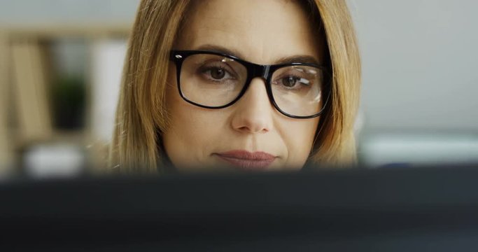 Close up of the Caucasian pretty blond woman in glasses sitting in front of the computer screen while working in the office.