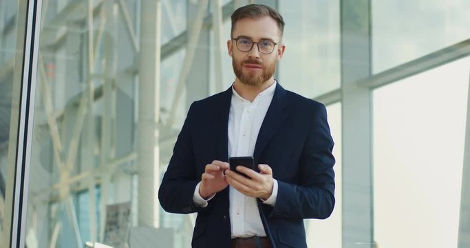 Portrait shot of the young handsome businessman taping and texting on the smartphone in his hands, then rising head and smiling to the camera.