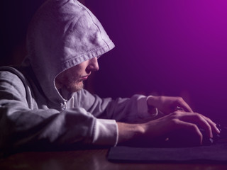 portrait of hooded man typing text on keyboard in front of pc monitor late in the night