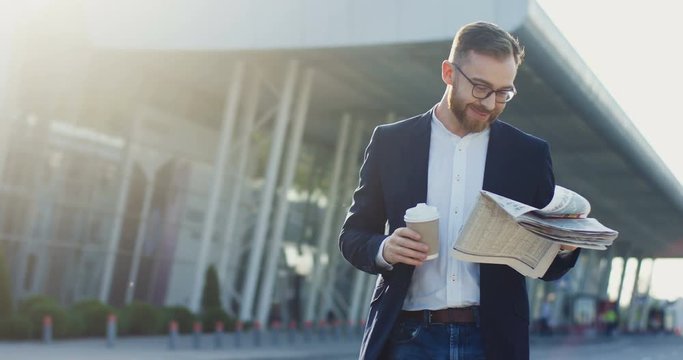 Young handsome Caucasian businessman walking near the big modern airport, drinking coffee and reading a newspaper.