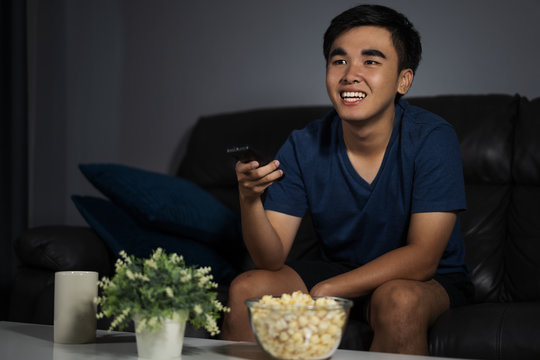 cheerful man holding remote control and watching TV while sitting on sofa at night
