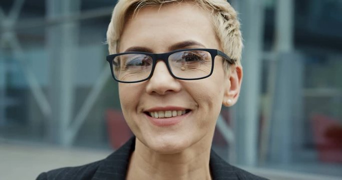 Close up of the charming Caucasian woman in glasses with short hair looking straight into the camera on the modern glass building background. Portrait.