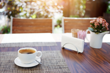 White Cup of black coffee with a saucer on a linen napkin and a vase with pink flowers on a table. Front view.