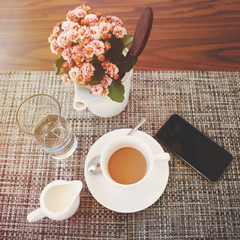 White cup of black coffee with a saucer, a vase with pink flowers, a glass with water, a creamer...