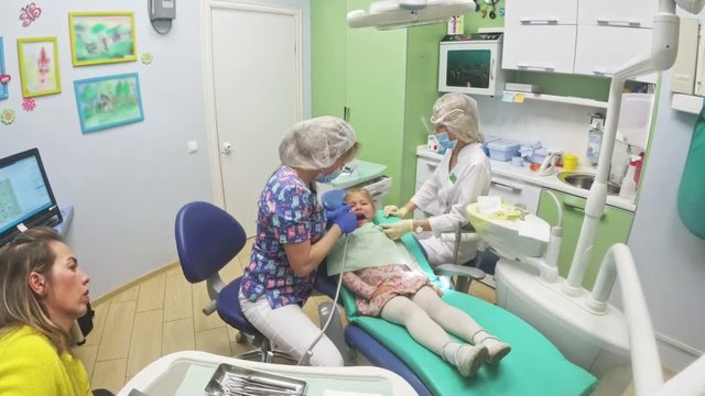 Child with a mother at a dentist's reception. The girl lies in the chair, behind her mother. The doctor works with an assistant. Procedure for drilling a tooth. Setting a temporary seal. Using the x