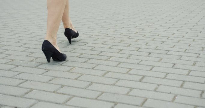 Rear of the female legs in the short skirt and shoes with heels walking on the street.