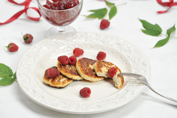 On a plate of syrniki with fresh berries of raspberries. Next to a bowl of raspberry jam. The leaves of raspberry and ribbon of crimson color complement the composition. Light background. Close-up.