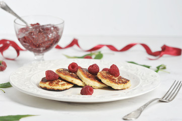 Cheesecakes with raspberries on a plate.  Next to the plate is a fork and a bowl with raspberry jam. A light background is complemented by raspberry leaves and a ribbon of crimson color. Close-up.
