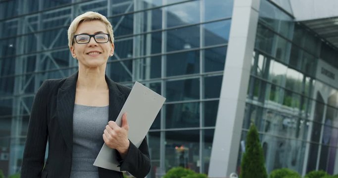 Portrait shot of the Caucasian beautiful woman office worker standing near big glass office center with documents in hands and smiling to the camera.