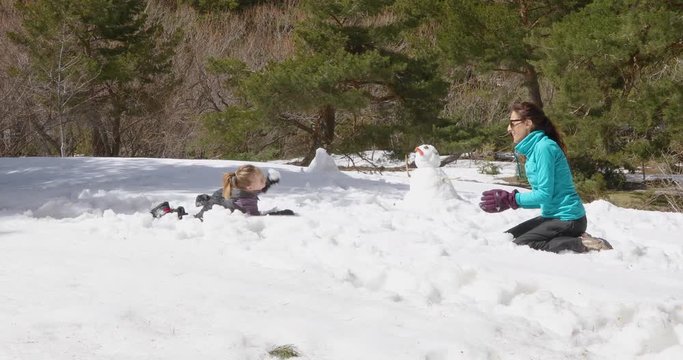 Four years old blonde child, and woman mother, sitting on snow in winter mountain, next to snowman, playing to throw snowballs. 4k video. 
