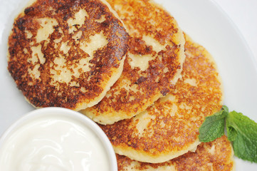 Cheesecakes on a white plate. Next to the cheesecakes is a cup of sour cream. Syrniki are decorated with a leaf of fresh mint. View from above. Close-up. Macro photography.