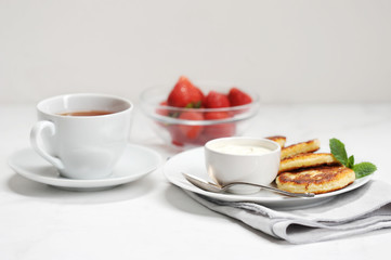 Table setting for breakfast. On a plate syrniki with sour cream. Next a cup of tea and a bowl of fresh strawberries. Light background. Close-up.