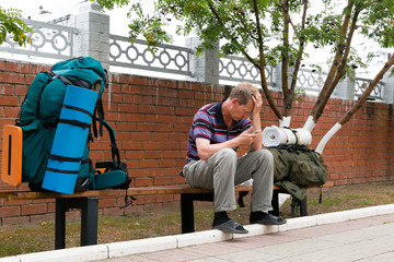 A man is sitting on a bench with large backpacks. A traveler is sitting with his bags at the railway station with a phone in his hand.