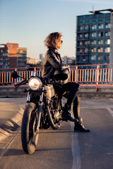Sexy biker beautiful female in black trendy leather jacket sit on vintage custom made caferacer motorcycle. Urban roof parking, sunset in big city. Traveling and active hipster lifestyle. Girls power.
