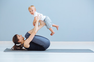 Sport, motherhood and active lifestyle concept - side view of mother workout together with her toddler boy over gray wall background. Mother having fun and playing with her little son.