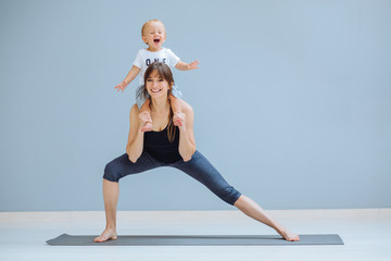 European sportive mother piggybacking her toddler baby son in fitness clothing on gray background. Lounge exercise. Motherhood, healthy lifestyle concept.