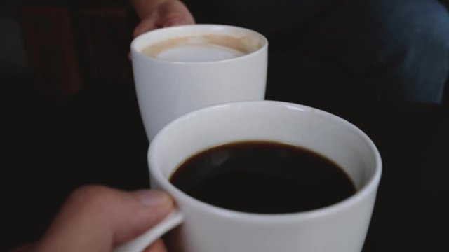 Close up image of two people clinking white coffee mugs in cafe