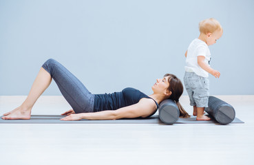 Young mother relaxing her neck and back with foam roller does physical pilates exercises while her toddler baby son playing near. Fitness, happy maternity sport with children concept.