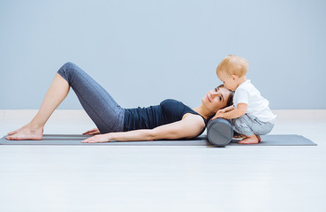 Young mother relaxing her neck and back with foam roller does physical pilates exercises while her toddler baby son playing near. Fitness, happy maternity sport with children concept.