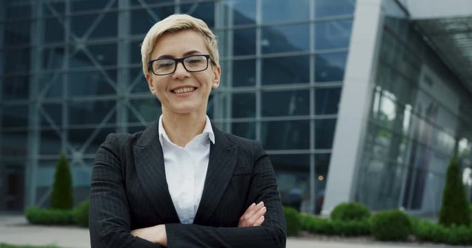 Cheerful Caucasian woman in the glasses and with short blond hair looking at the side and then turning her head to the camera with a smile on the big glass building background.
