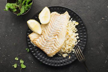 A modern dish is sea food. Fried cod white fish with rice on a side dish with slices of juicy lemon on a black plate and a black background with micro greenery. Top View.