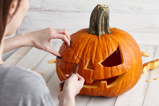 Woman carving big orange pumpkin into jack-o-lantern for Halloween holiday decoration on white wooden planks, close up view