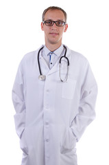 Adult doctor standing in white lab coat with hands in pocket with glasses and stethoscope and smiling on white background