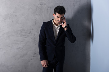 A serious handsome young businessman in a black suit talking on his phone in front of a grey wall in a studio.