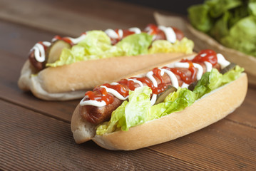 Two homemade Hot Dogs with mayonnaise, ketchup, and green lettuce leaves decorated with baking paper, over wooden table.