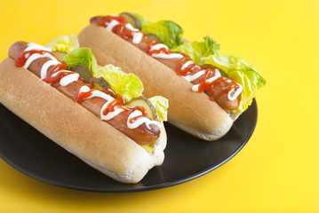 Two homemade Hot Dogs with mayonnaise, ketchup, and green lettuce leaves in black plate over yellow background. Top view.