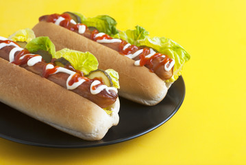 Two homemade Hot Dogs with mayonnaise, ketchup, and green lettuce leaves in black plate over yellow background. Top view.