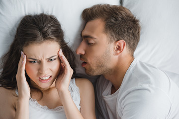 young angry woman having headache while lying with snoring husband in bed