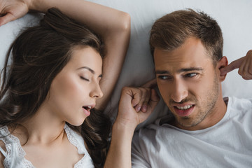 Fototapeta na wymiar top view of man closing ears and looking at snoring wife in bed
