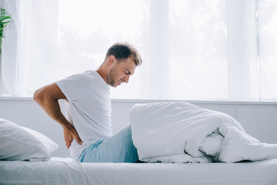 Side View Of Young Man In Pajamas Sitting On Bed And Suffering From Back Pain