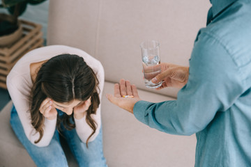 cropped shot of man holding glass of water and pills while woman suffering from headache