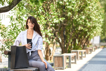 Pretty brunette drinking coffee sitting on wooden bench near paper bags.