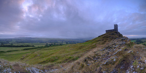 Fototapeta na wymiar Summer sunrise on Brentor showning St Michael's church atop the tor with dramatic weather clouds of showers and mist, on the western edge of the Dartmoor National Park. UK