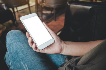 Mockup image of a woman's hand holding white mobile phone with blank desktop screen on thigh while sitting in cafe